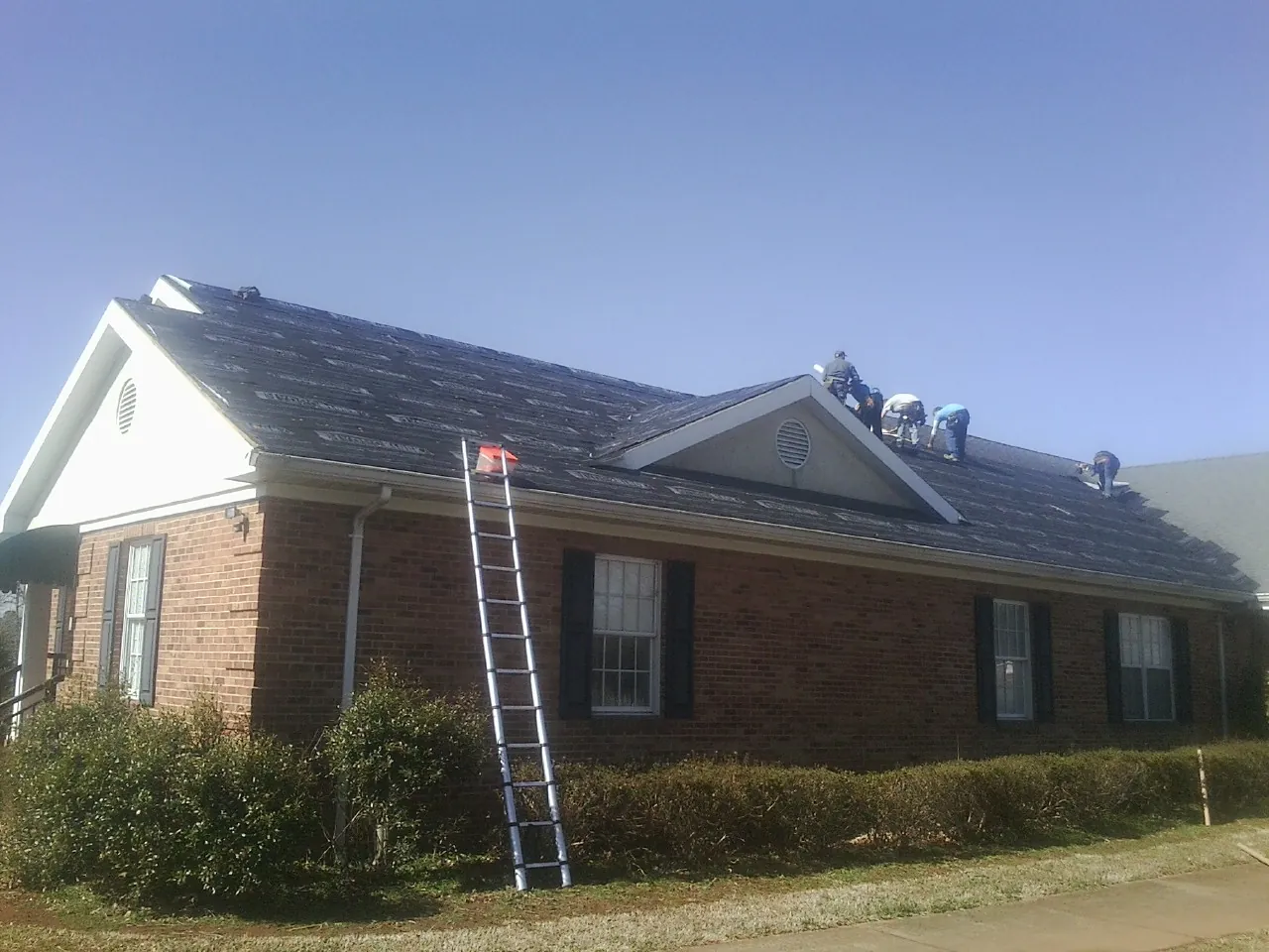 Crew tearing shingles off a brick ranch roof with a ladder set — Metal Roof Repair work by our crew in Newton